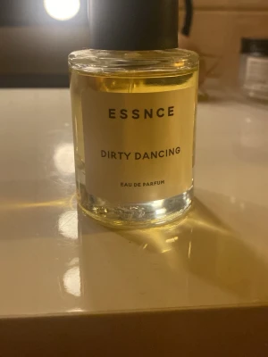 ESSNCE Dirty Dancing Eau de Parfum - En stilren parfymflaska från ESSNCE med doften Dirty Dancing. Flaskan är cylindrisk i genomskinligt glas med svart lock och en enkel, modern etikett. Innehållet är ljusgult och ger ett exklusivt intryck.