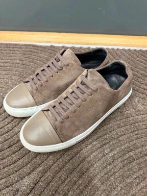 Axel Arigato skor  - Säljer ett par stilrena sneakers från Axel Arigato i brunt mocka med matchande bruna skosnören och tåparti i slätare läder. Skorna har vit sula och diskret guldfärgad logga på sidan. Perfekta för dig som gillar minimalistisk och clean stil.