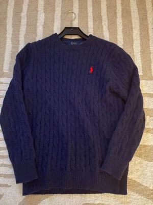 Mörkblå kabelstickad tröja Polo Ralph Lauren - Kabelstickad mörkblå tröja från Polo Ralph Lauren med rund halsringning och röd broderad logga på bröstet. Tröjan har långa ärmar och ribbade muddar vid ärmslut och nederkant. Perfekt för dig som gillar klassisk och stilren look.