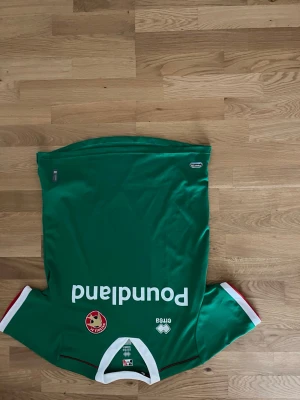 Grön fotbollströja med vita detaljer - Säljer en grön fotbollströja med vita och röda detaljer vid ärmsluten och halsen. Tröjan har tryck med texten 'Poundland' och klubbmärke på bröstet. Tillverkad i ett lätt och snabbtorkande material, perfekt för träning eller match.