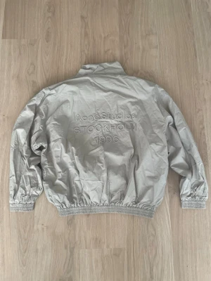 Grå vindjacka från Acne Studios - Snygg grå vindjacka från Acne Studios med broderad text 'Acne Studios STOCKHOLM 1996' på ryggen. Jackan har dragkedja framtill, elastiska muddar vid ärmslut och midja samt en hög krage. Tillverkad i ett lätt och prassligt material.