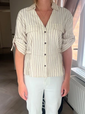 Randig beige skjorta med knappar - Säljer en beige skjorta med tunna vertikala ränder i mörkrött. Skjortan har knappar framtill, två bröstfickor och uppvikta ärmar med knappdetalj. Passformen är normal och materialet känns som bomull. Perfekt till jeans eller kjol!