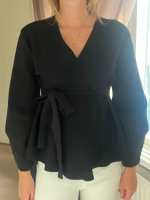 Svart omlottblus med knytband - Stilren svart omlottblus med långa ärmar och knytband i midjan. Blusen har en v-ringad hals och ett lätt utsvängt fall nedtill. Perfekt för en trendig och enkel look.