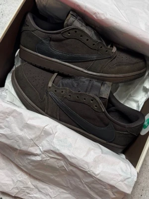 Bruna låga sneakers med svart swoosh - Air Jordan 1 Retro Low OG SP Travis Scott Velvet Brown. Låg modell med perforerad tåbox och klassisk snörning. Snygg och stilren design som passar till det mesta. Skorna har en mörkbrun yttersula och diskreta sömmar. Är tillgängliga i alla storlekar 