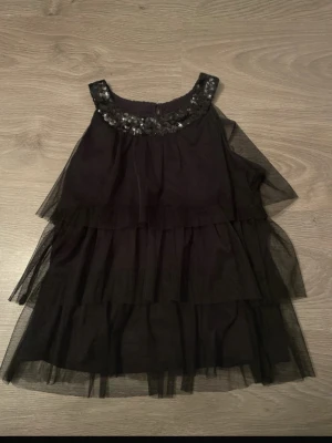 Svart volangklänning med paljetter - Säljer en svart klänning med flera lager volang i tunn tyll och paljetter vid halsringningen. Klänningen är ärmlös och har en luftig, A-linjeformad siluett som ger en elegant look.