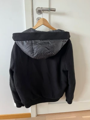 Svart cardigan från Bvaldi - Svart pufferjacka från Bvaldi med huva och quiltad front. Jackan har ribbade muddar, dragkedja framtill och två fickor med dragkedja. Ärmarna är stickade för extra komfort och jackan har en patch på ena ärmen. Perfekt för kalla dagar.