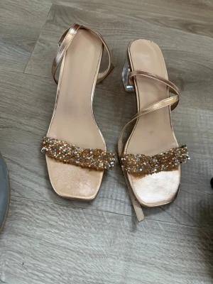 Glittriga guldiga pumps med remmar - Säljer ett par eleganta pumps i guld med fyrkantig tå och transparent klack. Skorna har tunna remmar runt ankeln och en bred rem framtill dekorerad med glittrande stenar. Perfekta för att lyfta vilken outfit som helst!
