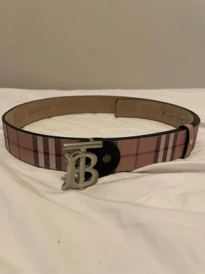 Rutig Burberry bälte med silverdetalj - Säljer ett klassiskt Burberry bälte med rutigt mönster i rosa, beige och svart. Bältet har ett stort silvrigt 'B'-spänne och är gjort i skinn. Perfekt accessoar för att lyfta din outfit.
