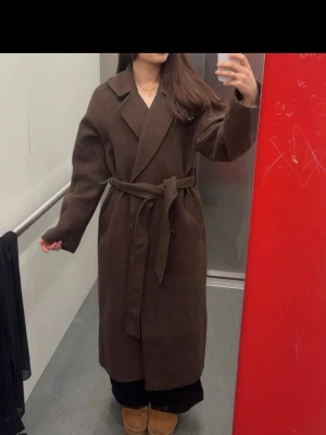 Slutsåld  kappa NA-KD - Stilren mörkbrun kappa i lång modell med breda slag och knytskärp i midjan. Kappan har en rak passform och är tillverkad i ett tjockare tyg som ger en lyxig känsla. Perfekt för kalla dagar och ger en snygg siluett. Storlek S
