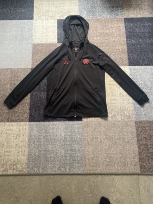 Svart PSG x Jordan zip hoodie - Säljer en svart zip hoodie från Jordan i samarbete med PSG. Hoodien har röda loggor på bröstet, hel dragkedja och två fickor framtill. Tillverkad i ett mjukt och sportigt material, perfekt för en avslappnad streetwear-look.