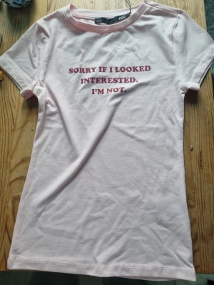 Ljusrosa t-shirt med texttryck - Trendig ljusrosa t-shirt från H&M med röd text på bröstet: 'SORRY IF I LOOKED INTERESTED. I'M NOT.' Klassisk rund halsringning och korta ärmar. Perfekt för dig som gillar statement-plagg och enkel stil.