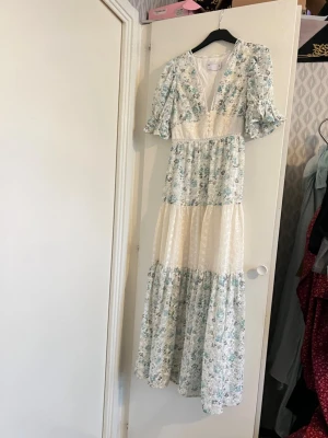 Blommig långklänning med volang - Romantisk långklänning med blommigt mönster i ljusblått och vitt. Klänningen har v-ringning, volangdetaljer och korta ärmar. Tillverkad i ett lätt och luftigt material, perfekt för varma dagar. Passformen är normal och klänningen har en feminin siluett.