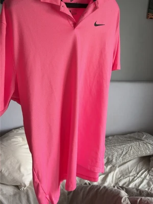 Rosa nike golf pikétröja  - Snygg rosa pikétröja från Nike med klassisk krage och korta ärmar. Tröjan har en enkel design med Nike-logga på bröstet och är tillverkad i ett mjukt och bekvämt material. Perfekt för en sportig och avslappnad stil.