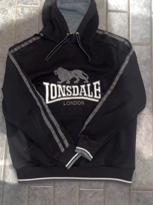 Svart Lonsdale hoodie med lejon - Svart hoodie från Lonsdale med stor logga och lejon på bröstet. Har huva med dragsko, gråa detaljer längs ärmarna och ribbade muddar med vit rand. Mjuk insida och klassisk känguruficka framtill. Perfekt för en chill och sportig look.
