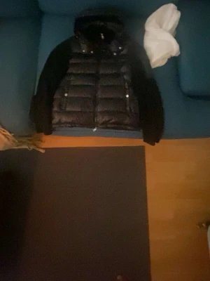 Svart dunväst med huva - Snygg svart dunväst med quiltad framsida och huva. Västen har dragkedja framtill och två fickor med knappar. Perfekt för lager-på-lager och ger en sportig look. Materialet är glansigt på framsidan och huvan, medan ryggen och ärmarna är i mjukare tyg.