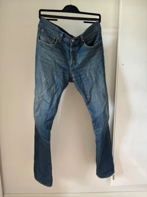 Levi’s 501 - Säljer ett par klassiska blå jeans med rak passform och fem fickor. Jeansen har knappgylf och tydliga kontrastsömmar. Materialet är slitstark denim i bomull och färgen är mellanblå med lätt tvättade detaljer. Perfekta för en avslappnad stil.