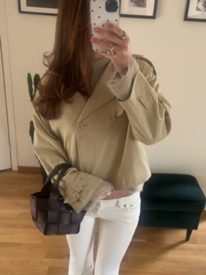 Kort beige trenchcoat med dubbelknäppning - Snygg kort trenchcoat i beige med dubbelknäppning framtill och axelklaffar. Jackan har bälte vid ärmsluten (en är borta) och en klassisk krage. Perfekt för dig som vill ha en trendig och stilren look. 