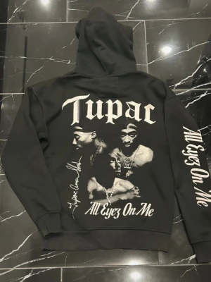 Svart Tupac hoodie All Eyez On Me - Svart hoodie med stor Tupac-print på ryggen och texten 'All Eyez On Me' på både ryggen och ena ärmen. Framsidan har Tupac-logga i vitt. Klassisk huva med dragsko och känguruficka. Materialet är mjukt och bomullsliknande.