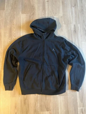 Svart zip hoodie från Jordan - Säljer en svart hoodie från Jordan med dragkedja framtill och två stora fickor. Hoodien har en liten Jumpman-logga på bröstet och är tillverkad i mjukt bomullsmaterial. Perfekt för en avslappnad och sportig stil.