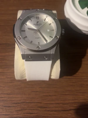 Klocka - Snygg klocka med silverfärgat ur och vit rem. Klockan har en stilren design med markerade skruvar på boetten och datumvisning. Quartz-urverk och tydliga index. Perfekt accessoar för dig som gillar exklusiv och modern stil. Priset kan diskuteras!