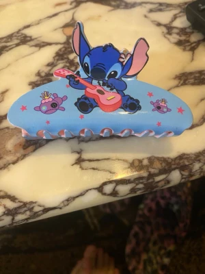 Blå hårklämma med Stitch-motiv - Söt blå hårklämma med motiv av Stitch som spelar gitarr och små lila björnar med kronor. Klämman har detaljer i rosa och lila samt stjärnor. Perfekt för dig som gillar Disney och vill ha en färgglad accessoar i håret.