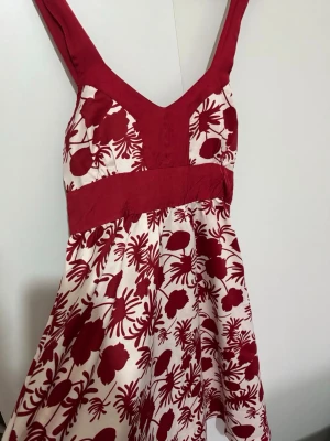 Rödvit blommig sommarklänning - Snygg kort röd och vit klänning med breda axelband och v-ringning. Klänningen har ett blommigt mönster och markerad midja, perfekt för varma dagar. Materialet känns lätt och luftigt, vilket gör den skön att bära under sommaren. Klänningen har inga skador och är oanvänd.