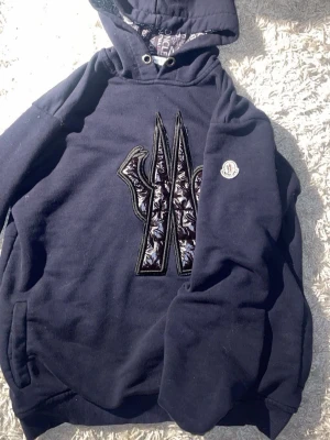 Svart hoodie från Moncler - Svart hoodie från Moncler med stort broderat M på bröstet och Moncler-logga på ärmen. Insidan av huvan har ett mönster med Moncler-tryck i vitt, rött och blått. Klassisk känguruficka och ribbade muddar.