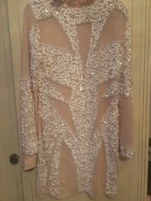Ljusrosa paljettklänning med pärlor - Säljer en unik ljusrosa kortklänning med långa ärmar, täckt av vita paljetter och pärldetaljer i ett geometriskt mönster. Klänningen har en elegant och iögonfallande design, perfekt för dig som vill sticka ut. Materialet känns mjukt och har en lätt transparent känsla.