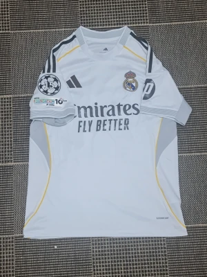 Real Madrid matchtröja Adidas vit - Snygg vit Real Madrid matchtröja från Adidas med klubbmärke, sponsortryck och Champions League-märke på ärmen. Tröjan har grå och gula detaljer samt 'AEROREADY'-material som andas. Perfekt för fotbollsträning eller att bära till vardags.