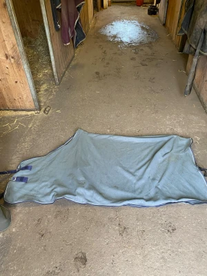 Grått hästtäcke med spännen - Säljer ett grått hästtäcke i fleece tyg med två spännen på ena sidan. Täcket är rektangulärt och har förstärkta kanter samt justerbara remmar. Perfekt för att hålla hästen varm i stallet. Med kryssgjordar, strl 125