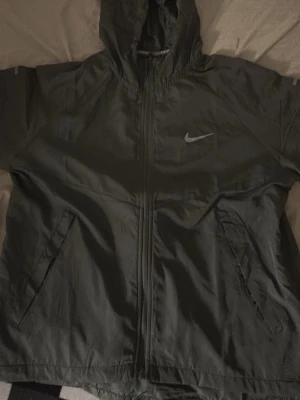 Grå Nike vindjacka med huva - Svart vindjacka från Nike med dragkedja framtill, två sidofickor och huva. Jackan har en minimalistisk design med Nike-logga på bröstet och är tillverkad i ett lätt, vindtätt material som passar perfekt för blåsiga dagar.
