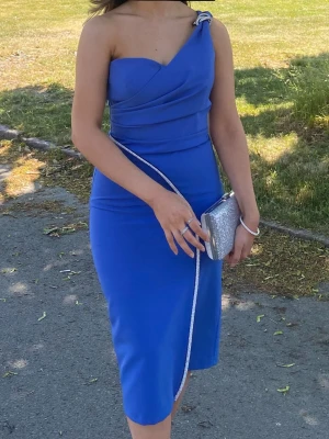 Blå one shoulder klänning - Snygg blå klänning med one shoulder-design och knytdetalj på axeln. Klänningen är figurnära och har en elegant, enkel siluett som passar perfekt till speciella tillfällen. Materialet känns mjukt och stretchigt för en bekväm passform. Köpt i en klänningsbutik i London. 