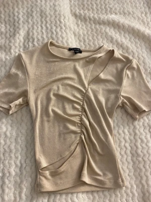 Beige omlottopp  - Trendig beige topp från AMISU med korta ärmar och snygg omlottdesign. Toppen har rynkade detaljer framtill och en asymmetrisk skärning som ger en cool look. Perfekt att styla med jeans eller kjol för en modern vibe.