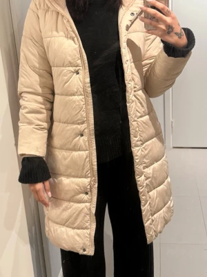 Beige lång dunjacka med huva - Snygg beige lång dunjacka med huva och tryckknappar framtill. Jackan har ett quiltat mönster och raka ärmar. Perfekt för kalla dagar och enkel att matcha med olika outfits. Jackan har en stilren look utan synliga loggor eller extra detaljer.