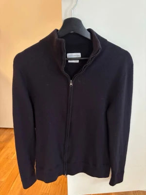 Svart merinoullskofta These Glory Days - Stilren svart kofta från These Glory Days i mjuk superior merinoull. Koftan har hel dragkedja framtill, hög krage och långa ärmar. Perfekt för lager på lager och enkel att matcha med olika outfits. Storlek M men mycket mer som en S.