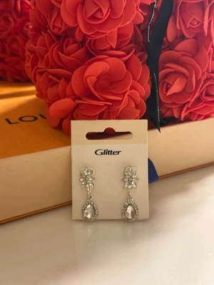 Örhängen Glitter - Säljer ett par eleganta silvriga örhängen från Glitter med gnistrande stenar i droppform och blommig detalj upptill. Örhängena har en klassisk och glammig look med små kristaller som ramar in den stora stenen. Perfekta för dig som gillar att sticka ut.