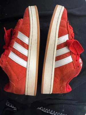 Röda Adidas sneakers med vita ränder - Snygga röda Adidas sneakers i mocka med klassiska vita ränder på sidorna och Adidas-logga på plösen. Skorna har röda skosnören och en vit/gummifärgad sula. Perfekta för dig som vill sticka ut med färg och sportig stil.