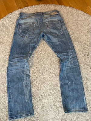 Blå raka jeans med slitningar - Säljer ett par blå diesel jeans med raka ben och tydliga slitningar för en avslappnad look. Jeansen har två bakfickor och klassisk femficksdesign. Materialet är denim i bomull och färgen är mellanblå med ljusare partier. Storlek 30/32.
