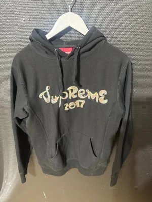 Svart Supreme hoodie 2017 - Svart hoodie från Supreme med broderad text 'Supreme 2017' i färgglada bokstäver på bröstet. Klassisk känguruficka och dragsko i huvan. Perfekt för dig som gillar streetwear och vill sticka ut med en limited edition piece.