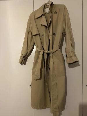 Beige klassisk trenchcoat med skärp från HM - Stilren beige trenchcoat med knytskärp i midjan, dubbelknäppning och dekorativa axelklaffar. Jackan har långa ärmar med justerbara remmar vid ärmsluten och klassisk krage. Perfekt för en trendig och tidlös look.