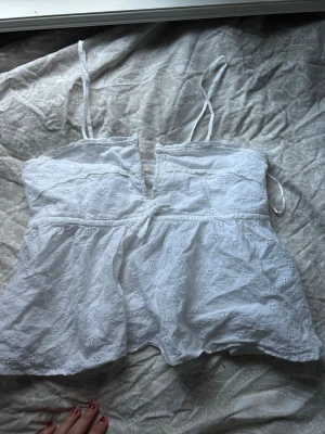 Vit broderad linnetopp Gina Tricot - Söt vit linnetopp från Gina Tricot med tunna axelband och broderade blommor. Toppen har en lätt v-ringning och ärmlös design, perfekt för varma dagar. Luftig och feminin modell som ger en somrig vibe.