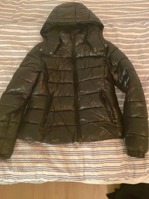 Svart pufferjacka med huva - Säljer en svart pufferjacka med stor huva och dragkedja framtill. Jackan har en glansig yta, breda quiltade paneler och två sidofickor med dragkedja. Perfekt för kalla vinterdagar och riktigt skön att bära. Materialet är troligtvis syntet.