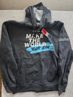 Mörkgrå hoodie från Clique x Celsius - Snygg mörkgrå hoodie från Clique i samarbete med Celsius. Tryck på bröstet med texten 'MAKE THE WORLD YOUR GYM' i vitt och blått samt Celsius-logga på ärmen. Klassisk känguruficka och dragsko i huvan. Mjuk insida och skön passform.