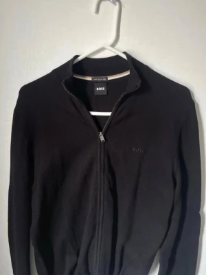 Ziptröja Hugo Boss - Snygg svart ziptröja från BOSS med diskret logga på bröstet. Tillverkad av italienskt garn och har ribbade muddar samt en stilren krage med beige och vit rand på insidan. Storlek S, inga defekter. Fraktar samma dag!