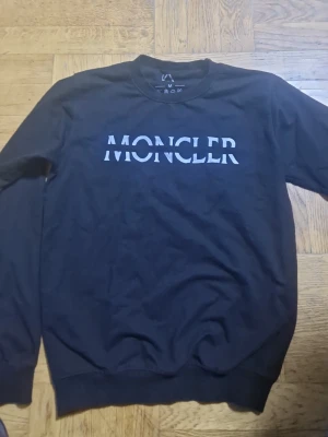 Svart sweatshirt från Moncler - Snygg svart sweatshirt från Moncler med stor vit logga tryckt över bröstet. Tröjan har rund halsringning, långa ärmar och ribbade muddar vid ärmslut och nederkant. Perfekt för en clean och stilren look.