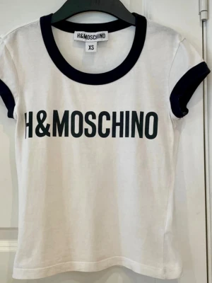 Vit H&MOSCHINO t-shirt XS - Vit t-shirt från samarbetet H&MOSCHINO med svart text framtill. T-shirten har svarta kontrastdetaljer vid krage och ärmslut samt en klassisk, tight passform. Tillverkad i mjuk bomull som känns skön mot huden. Perfekt för en trendig och avslappnad look. Litet hål i nedre kanten, se bild