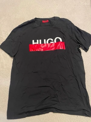 Svart t-shirt från Hugo med röd detalj - Cool svart t-shirt från Hugo med vit logotyp och en röd glansig detalj över bröstet. Klassisk passform och korta ärmar, perfekt för dig som gillar stilrena plagg med en twist. Materialet känns mjukt och bekvämt.
