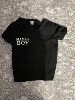 Svart set med text 'MAMA'S BOY' - Säljer ett svart tvådelat set från Shein med trycket 'MAMA'S BOY' i vitt på både t-shirt och shorts. Setet har enkel, rak passform och är gjort i mjukt material som känns bekvämt mot huden. Perfekt för dig som gillar statement-plagg och vill ha något avslappnat.