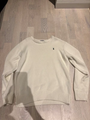 Vit stickad tröja från Polo Ralph Lauren - Klassisk vit stickad tröja från Polo Ralph Lauren med rund halsringning och ribbade muddar. Tröjan har den ikoniska lilla broderade loggan på bröstet. Perfekt för lager på lager och enkel att matcha med olika outfits.