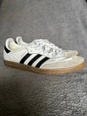 Adidas Samba OG – White / Black – Gum Sole - Klassiska Adidas Samba sneakers i vit / cream färg med svarta tre ränder och klassisk gum sole.  En ikonisk modell inspirerad av retro football och terrace culture, vilket gör dem väldigt populära igen.  Detaljer • Adidas Samba • White / black • Klassisk gum sole • Retro terrace style • Bekväma och slitstarka  Perfekta till jeans, cargos eller casual outfits.  📦 Skickas snabbt 💬 Hör av dig vid frågor